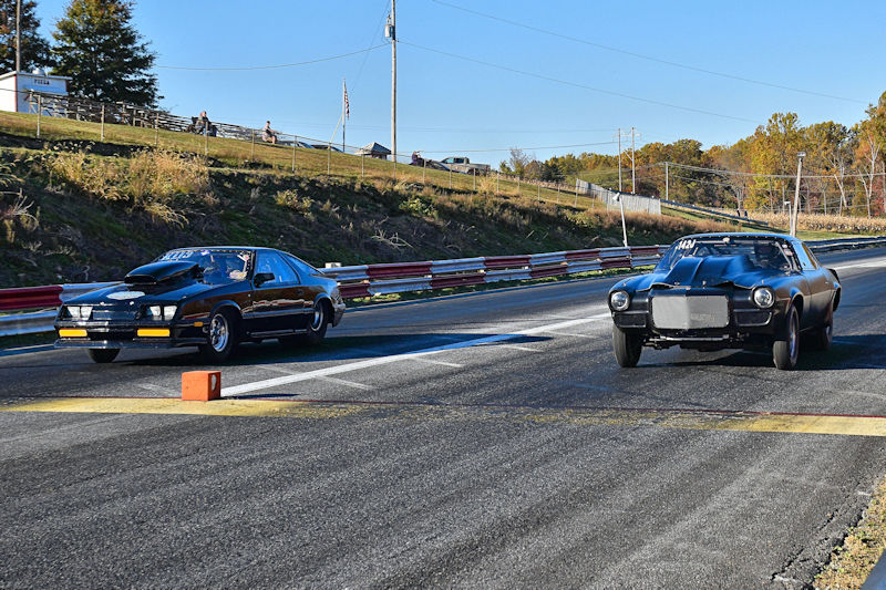 Two race cars competing on a drag strip, one black car on the left and another black car on the right, with spectators in the background.
