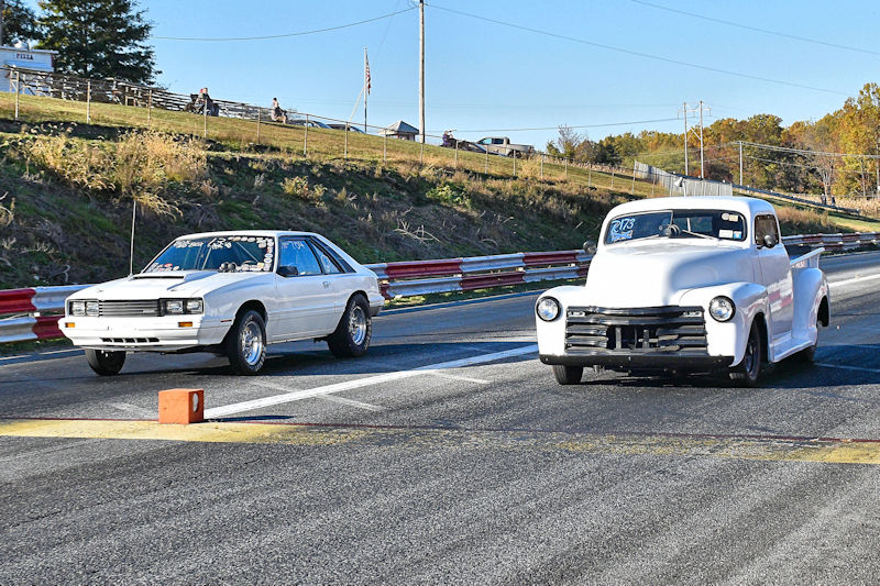 A white sports car and a white vintage pickup truck lined up at the starting line of a drag racing track, with spectators in the background.
