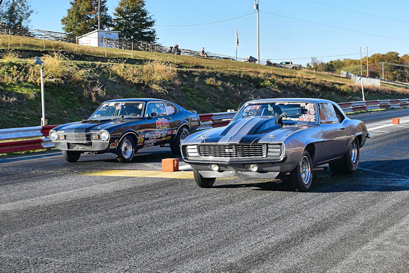 Two classic cars racing on a drag strip, one with a black exterior and the other silver with black stripes, surrounded by trees and a clear blue sky.