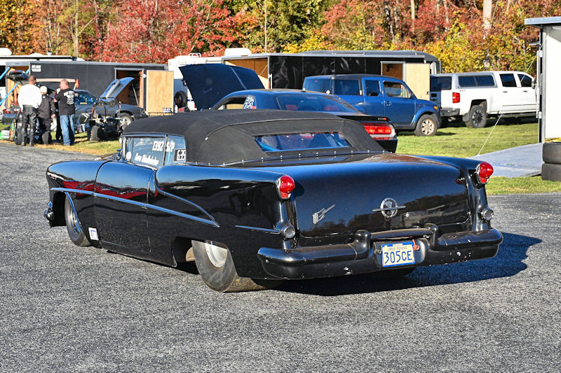 A classic black convertible car with a soft top and distinctive rear design, parked in a racing event setting surrounded by people and trailers.