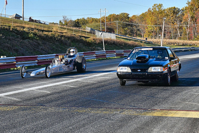 A drag race between a sleek silver dragster and a black muscle car on a racetrack, surrounded by autumn foliage.