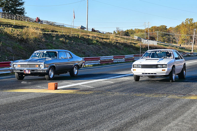 Two cars racing down a drag strip, one silver and one white, with spectators in the background on a sunny day.