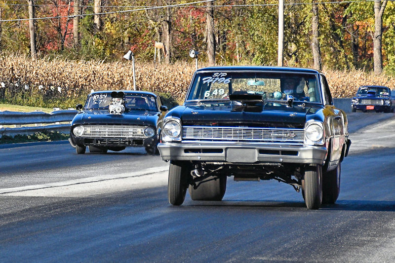 Two classic drag racing cars competing on a track, with one car lifting a front wheel off the ground.