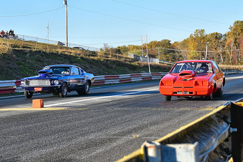 Two race cars competing on a drag strip, one blue with a hood scoop and the other red with a distinctive front end.