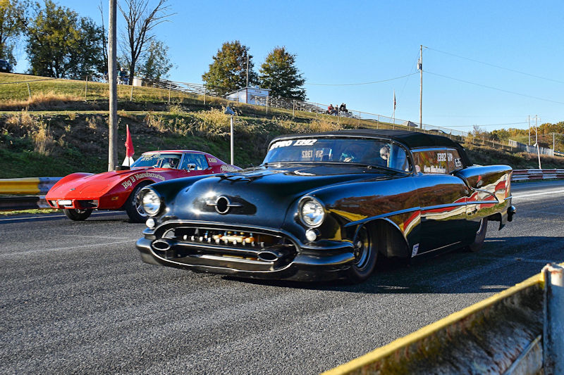 A black vintage car with unique features is racing on a track next to a red sports car, surrounded by trees in the background.