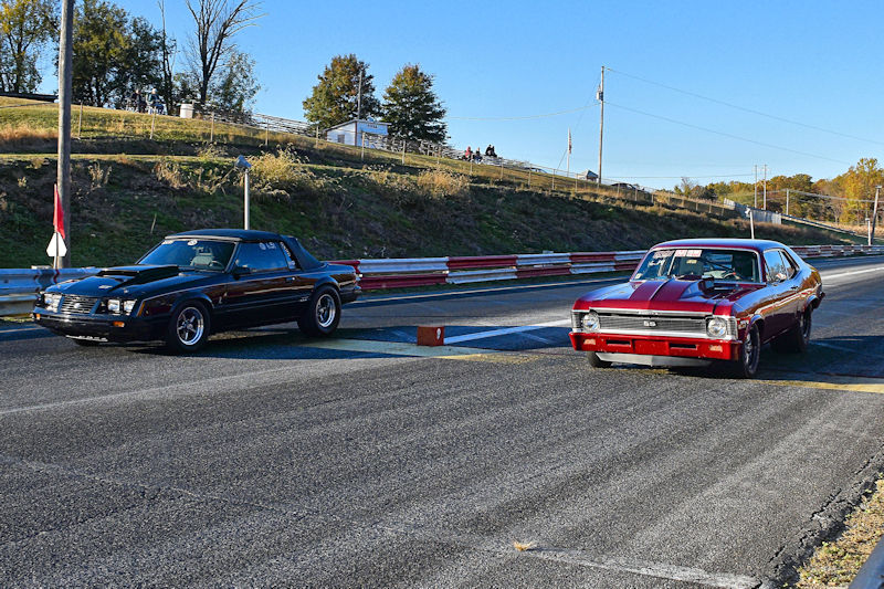 Two classic cars preparing for a race on a drag strip, one black and one red, with spectators in the background.