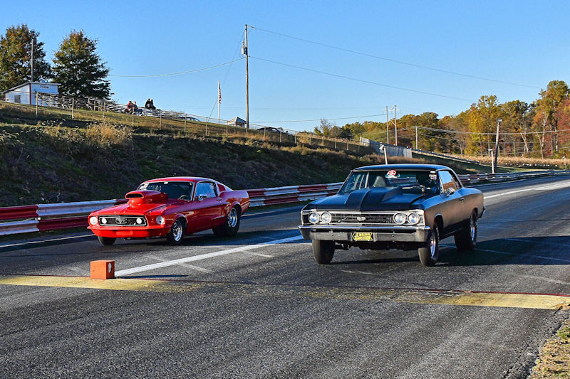 Two classic muscle cars, a red Mustang and a black Chevrolet, lined up at a drag strip ready for a race under clear blue skies.