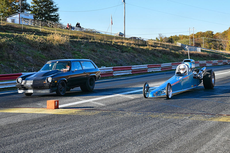 A black station wagon and a blue drag race car are lined up on a racing track, ready to compete. The background shows a hillside and spectators in the distance.