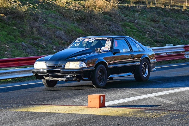 A black modified car racing on a track, showcasing a lowered stance and performance wheels, with colorful event decals on the windshield.