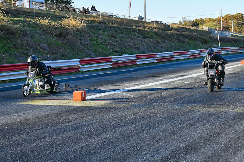 Two riders racing mini motorcycles on a drag strip, with one positioned closer to the starting line and the other further back.