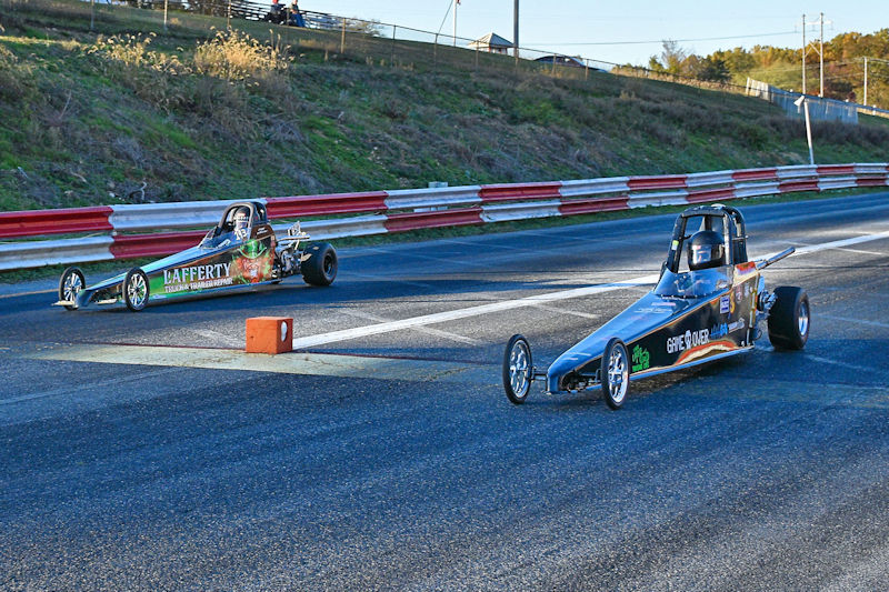 Two drag racing cars competing on a racetrack, with one car in the foreground and the other slightly behind. The cars are designed for racing, showcasing vibrant colors and sponsorship branding.