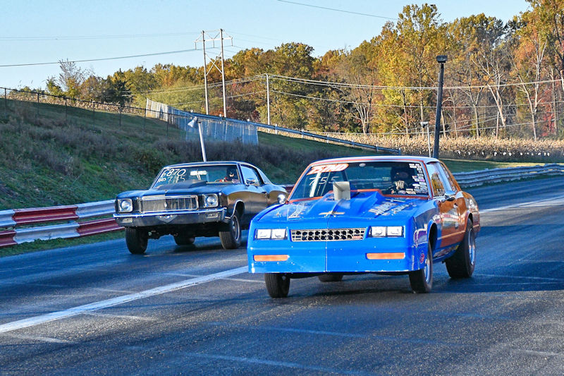 Two race cars on a drag strip, with one blue car leading ahead of a black car in the background.