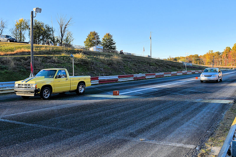 A yellow pickup truck and a silver sedan positioned on a drag strip, with a clear blue sky and autumn trees in the background.
