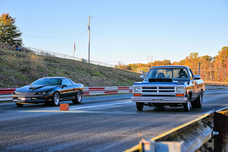 Two cars racing on a drag strip, with a black sports car on the left and a white pickup truck on the right.