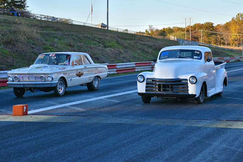 Two classic cars racing on a drag strip, one is a white pickup truck and the other is a vintage sedan, with a backdrop of trees and a blue sky.