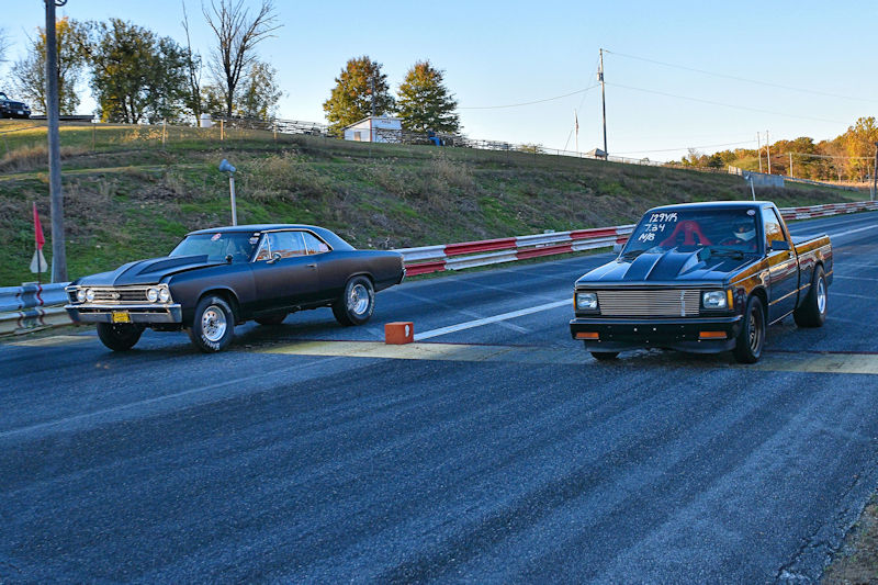Two cars positioned at the start line of a drag racing track, one a black classic muscle car and the other a modified pickup truck.