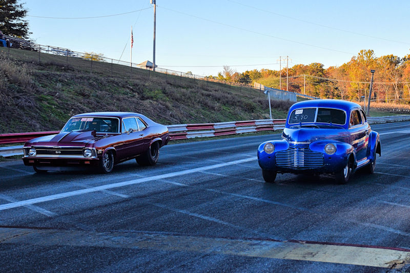 Two classic cars lined up on a racetrack: a maroon Chevrolet Nova and a blue vintage car, ready for a drag race at sunset.