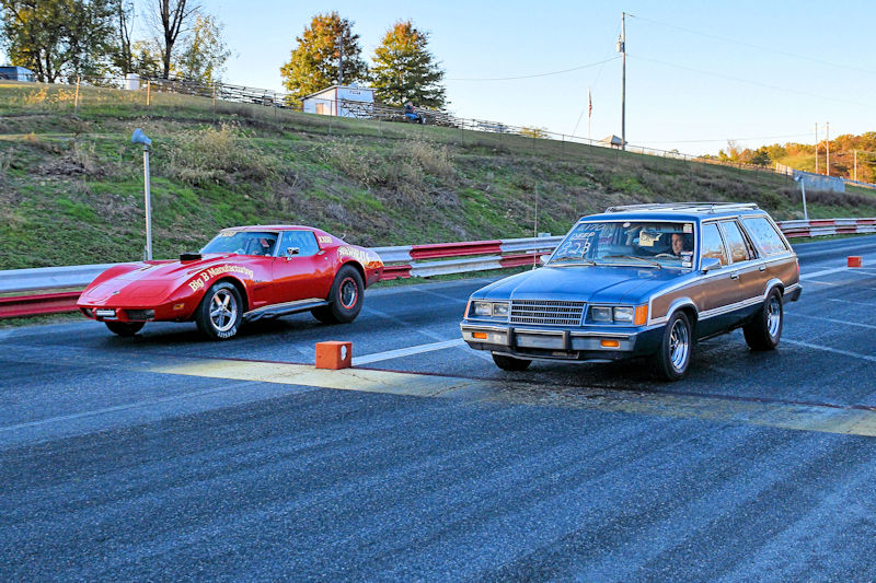 A red sports car and a brown station wagon lined up for a drag race on a racing strip.