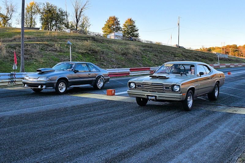 Two vintage cars lined up at a drag racing track, ready to race. The car on the left is a gray model with a rear spoiler, while the car on the right is a brown model with a distinctive hood scoop.