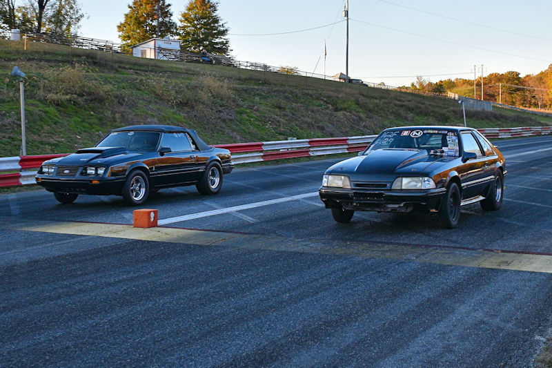 Two vintage cars lined up on a drag racing track, ready for a race. The car on the left has a convertible style with a black and brown color scheme, while the car on the right is a coupe with a black exterior. Both cars are positioned near the starting line.