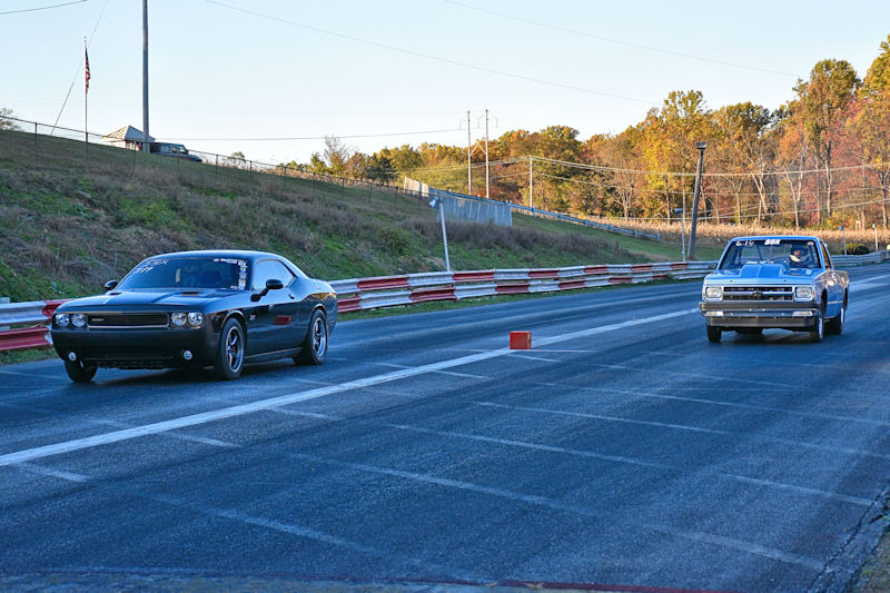 Two cars racing on a drag strip, with a black sports car on the left and a classic car on the right, showcasing a competitive scene.