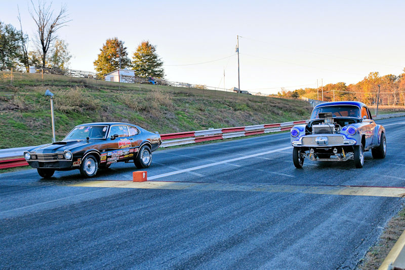 Two classic cars racing on a drag strip, one black and one blue, with trees and a fence in the background.