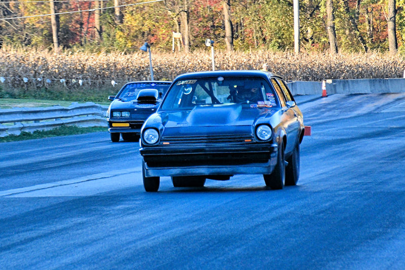 A black car racing on a drag strip, with a second car visible in the background. The scene features autumn foliage and a straight track.
