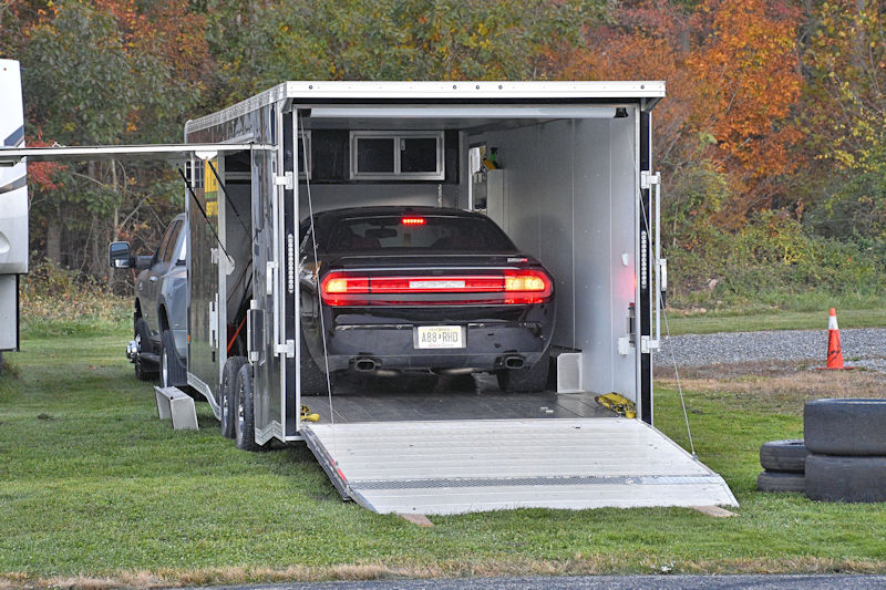 A black car being loaded into an open trailer with a ramp, set in a grassy area surrounded by autumn trees.