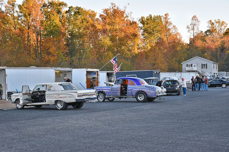 A parking area with two classic cars, a Mercury Comet and a purple vehicle, set against an autumn backdrop of colorful trees. People are gathered near the cars and trailers in the background.