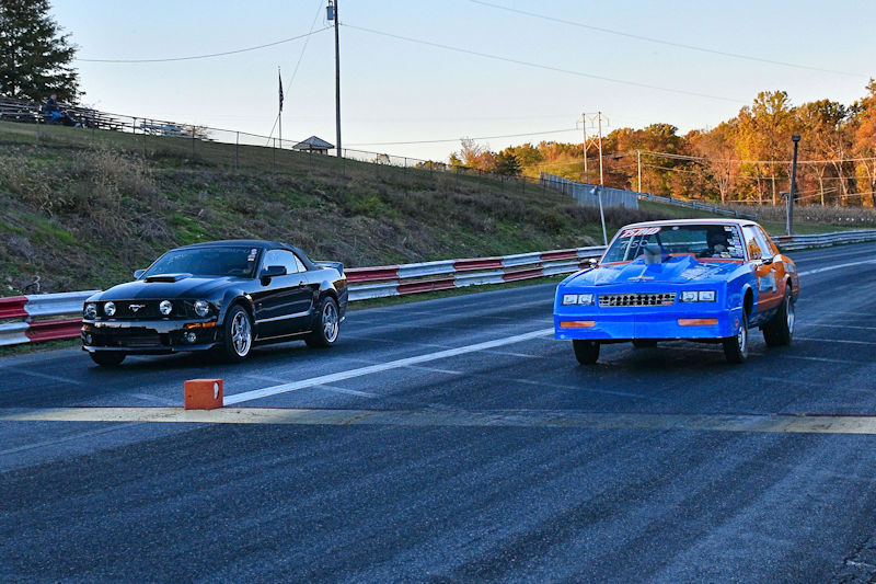Two cars racing on a drag strip, one black and one blue with orange accents.