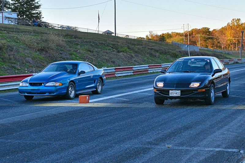 Two cars racing on a drag strip, one blue and one black, with a timing block in the center.