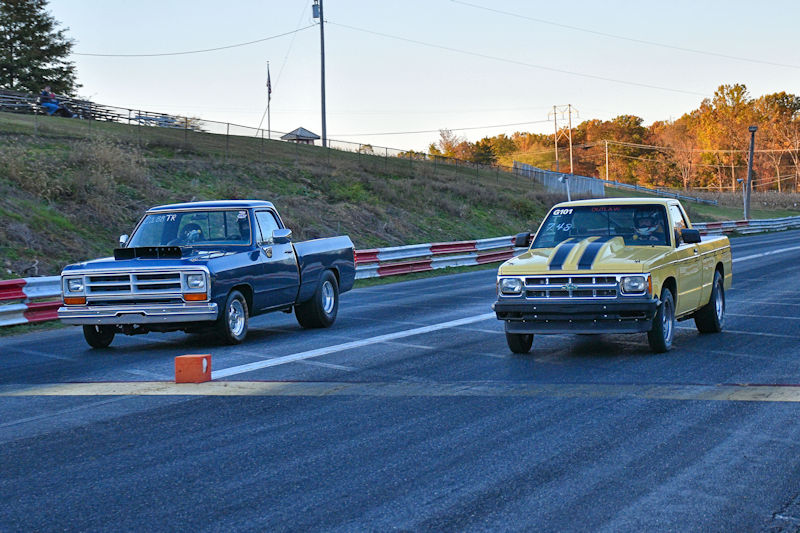 Two vintage pickup trucks racing on a drag strip, one blue and one yellow with black stripes.