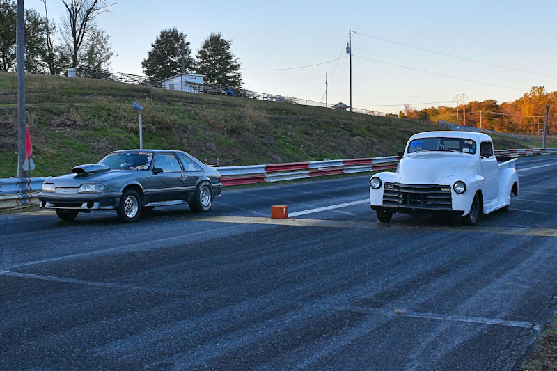 A gray muscle car and a classic white pickup truck are positioned at the starting line of a drag strip, ready for a race.