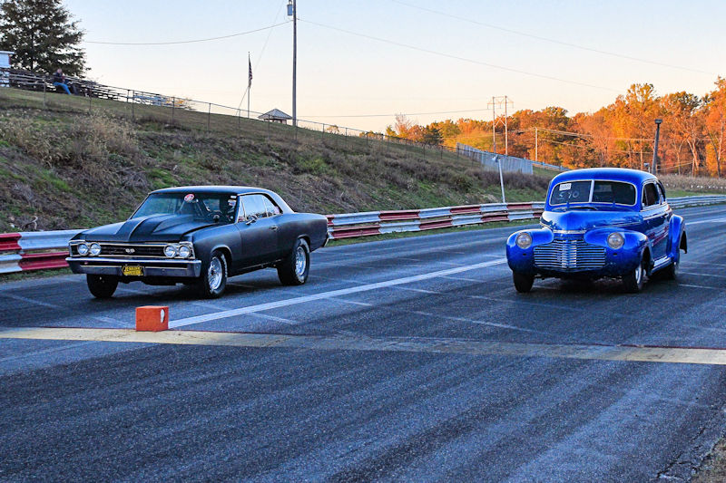 Two classic cars lined up for a race on a drag strip, with a dark-colored car on the left and a blue car on the right, set against a backdrop of trees and a clear sky.