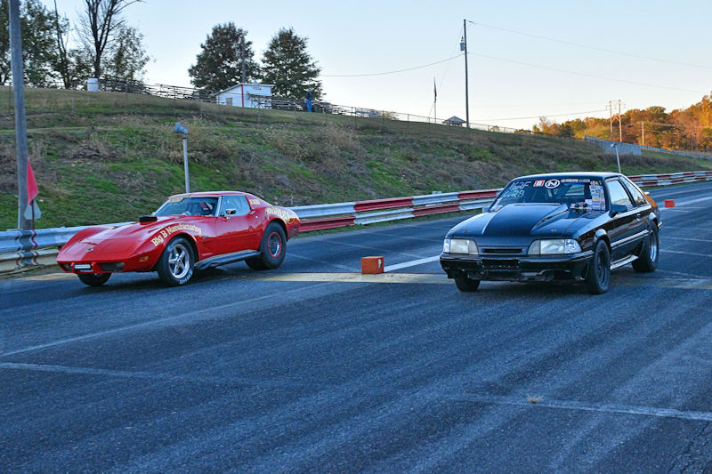 Two cars racing on a drag strip, a red sports car on the left and a black car on the right, with the track lined by barriers and trees in the background.