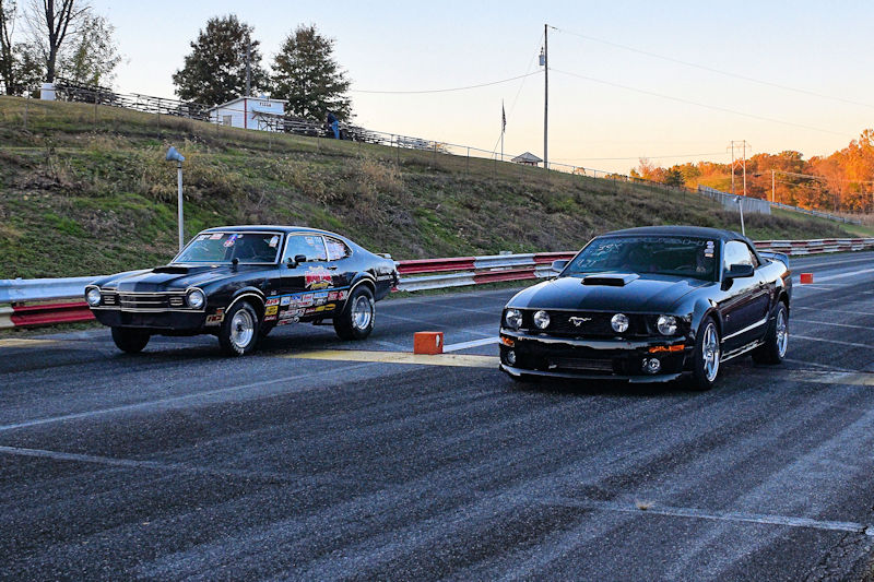 Two classic black cars lined up on a drag racing track, one is a vintage model and the other a modern Mustang, with an autumn landscape in the background.