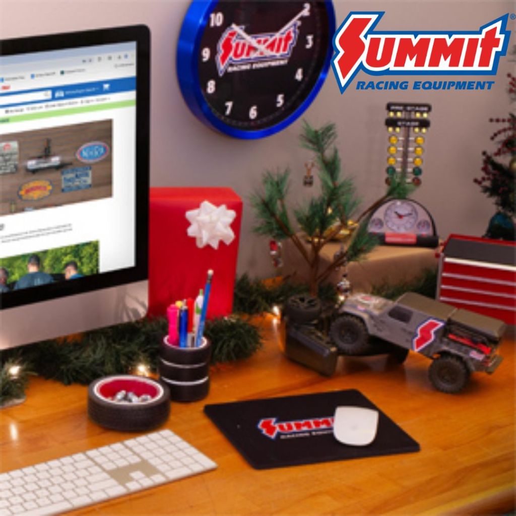 A workspace decorated with a festive theme featuring a computer displaying a website, a blue wall clock, a red gift box with a white bow, colorful pens in a cup, a miniature racing truck, and decorative greenery.